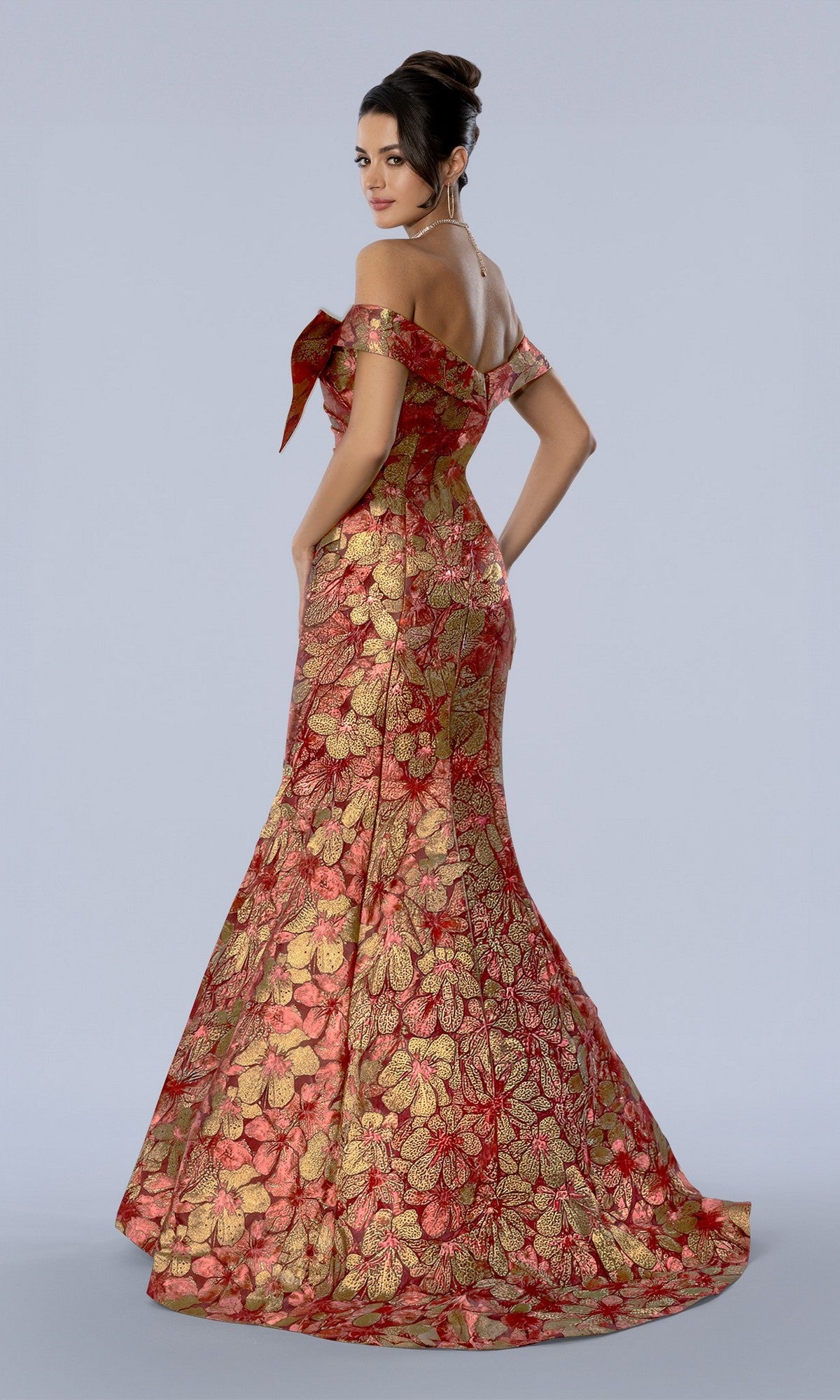 Woman in the Stella Couture 26354 long formal dress—an elegant, off-the-shoulder gown with a floor-length red and gold floral brocade design—stands facing slightly away against a plain light background.